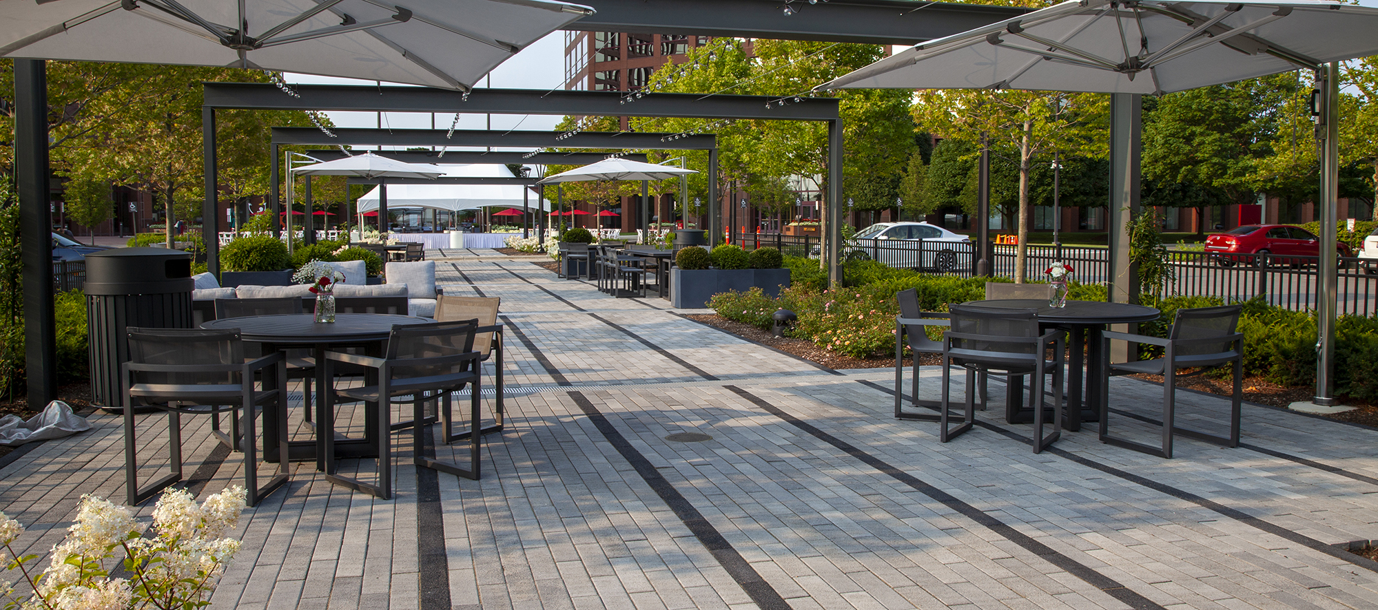 Two-toned Promenade Plank pavers delineate rows of walking spaces, with outdoor tables, chairs and umbrellas and landscaping on the sides.