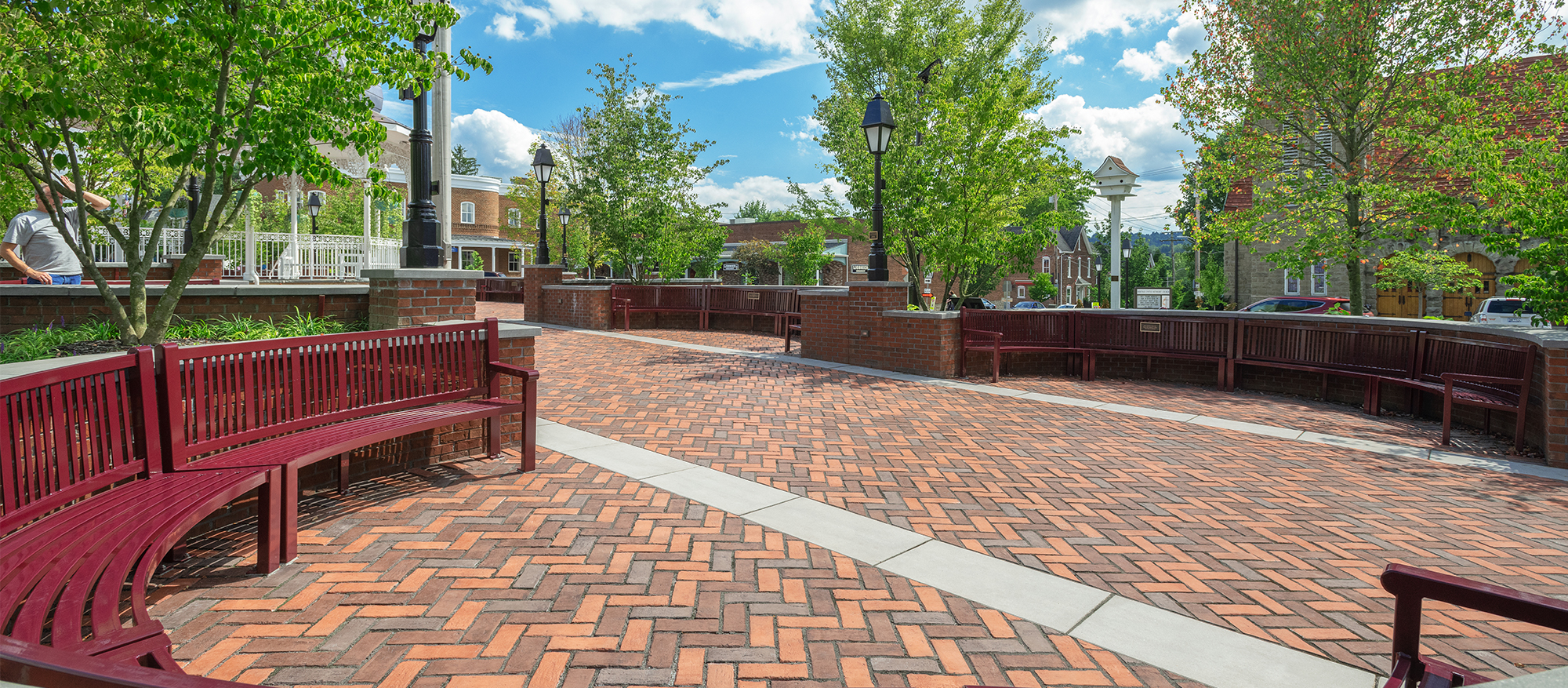 Rounded Burgundy benches in a tree-lined walled plaza made with Town Hall pavers in shades of red with gray slab stripes running through. 