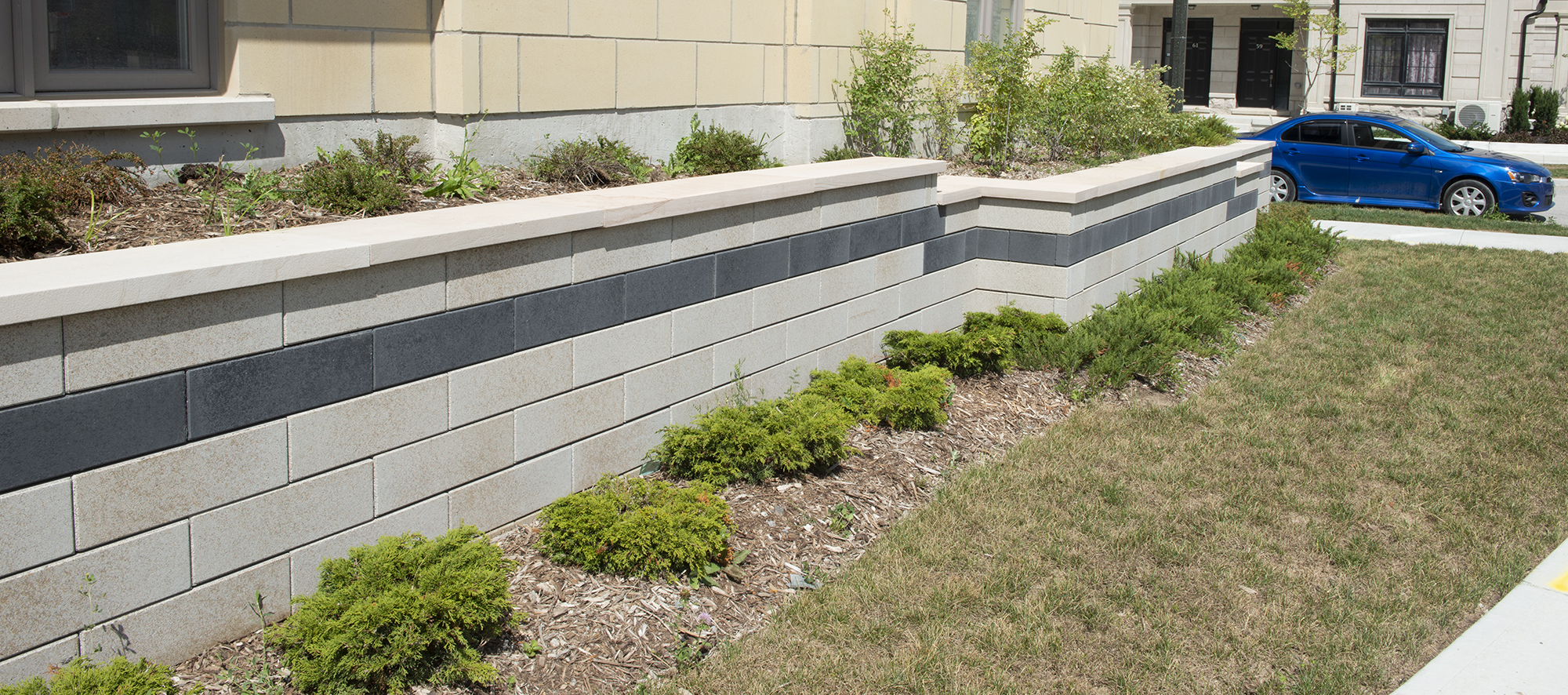 A two-toned U-Cara retaining wall provides multi-level garden beds featuring shrubbery along the side of the building.