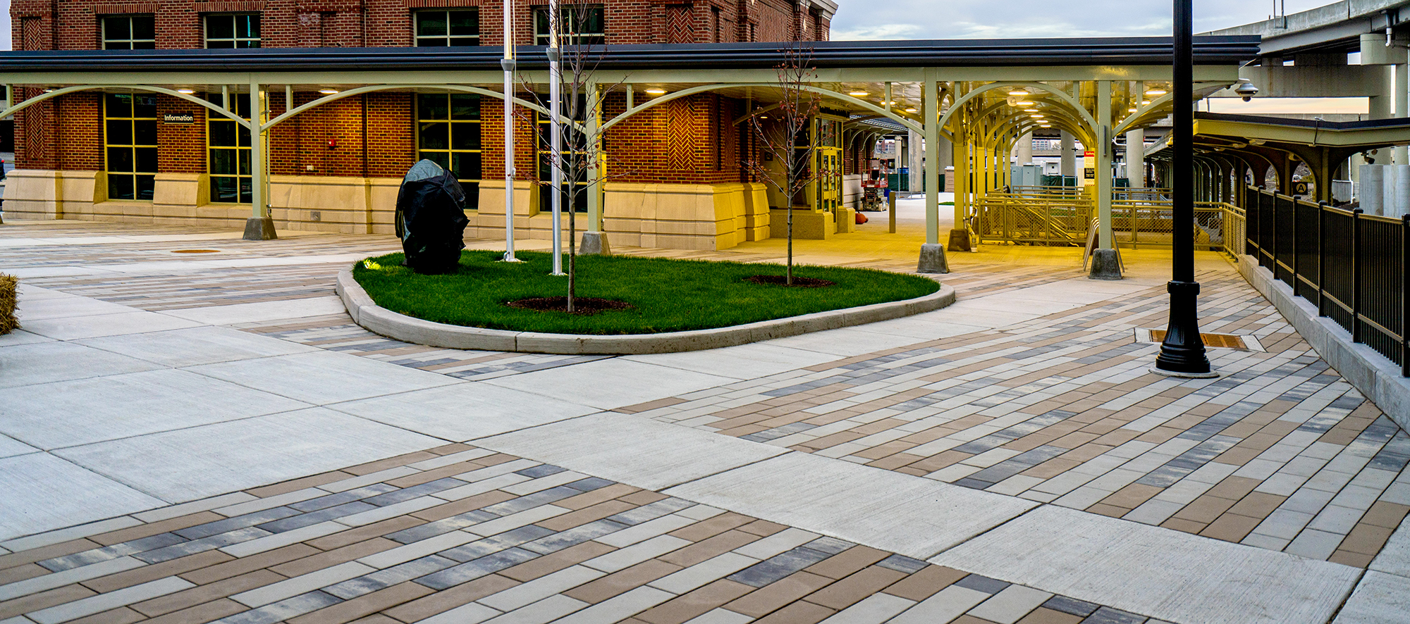 In front of a train station is a plaza in tri-color Promenade Plank pavers with concrete sidewalks and a small landscaped area.