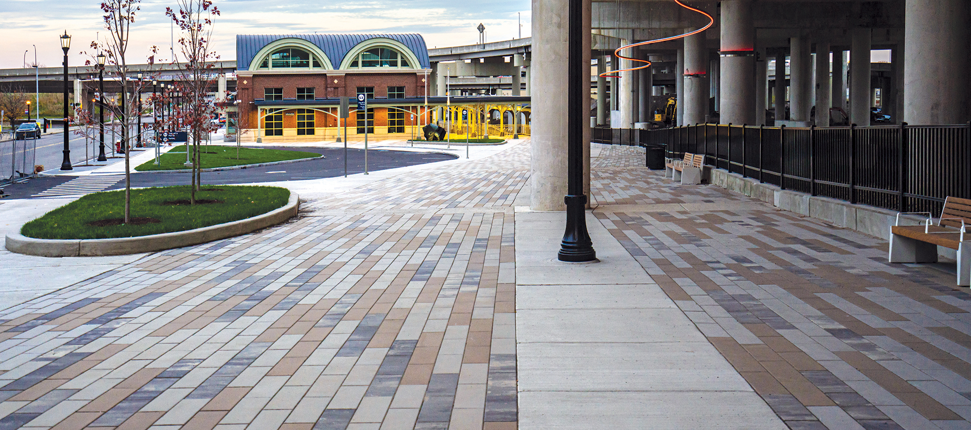 A pedestrian path made of tri-color Promenade Plank pavers, with landscaping and benches, runs alongside loading areas to a train station.