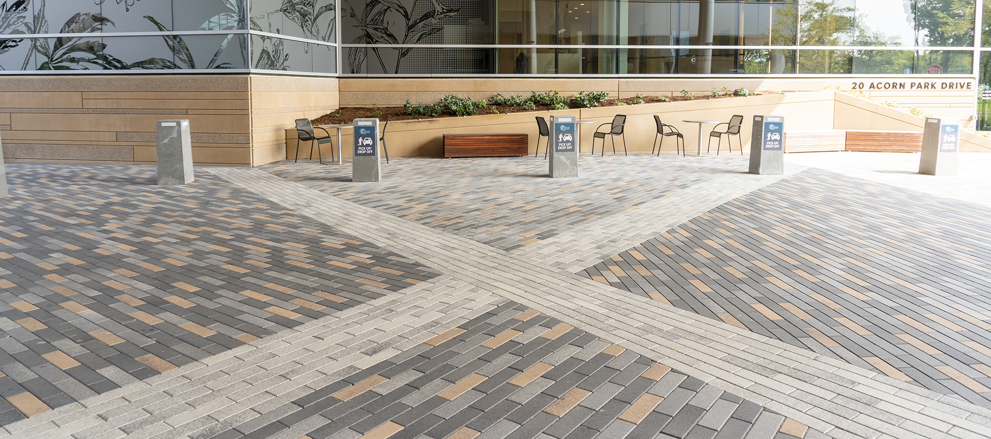 Promenade Plank pavers in three hues with a white X-shaped paver design cutting through the pattern, in front of a beige-grey building.