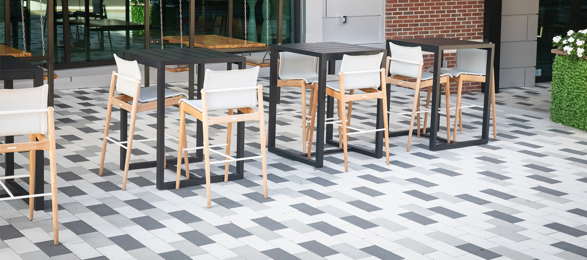Tri-colored Promenade Plank pavers form a checkboard pattern underneath a quartet of bar chairs and tables in front of a brick building.