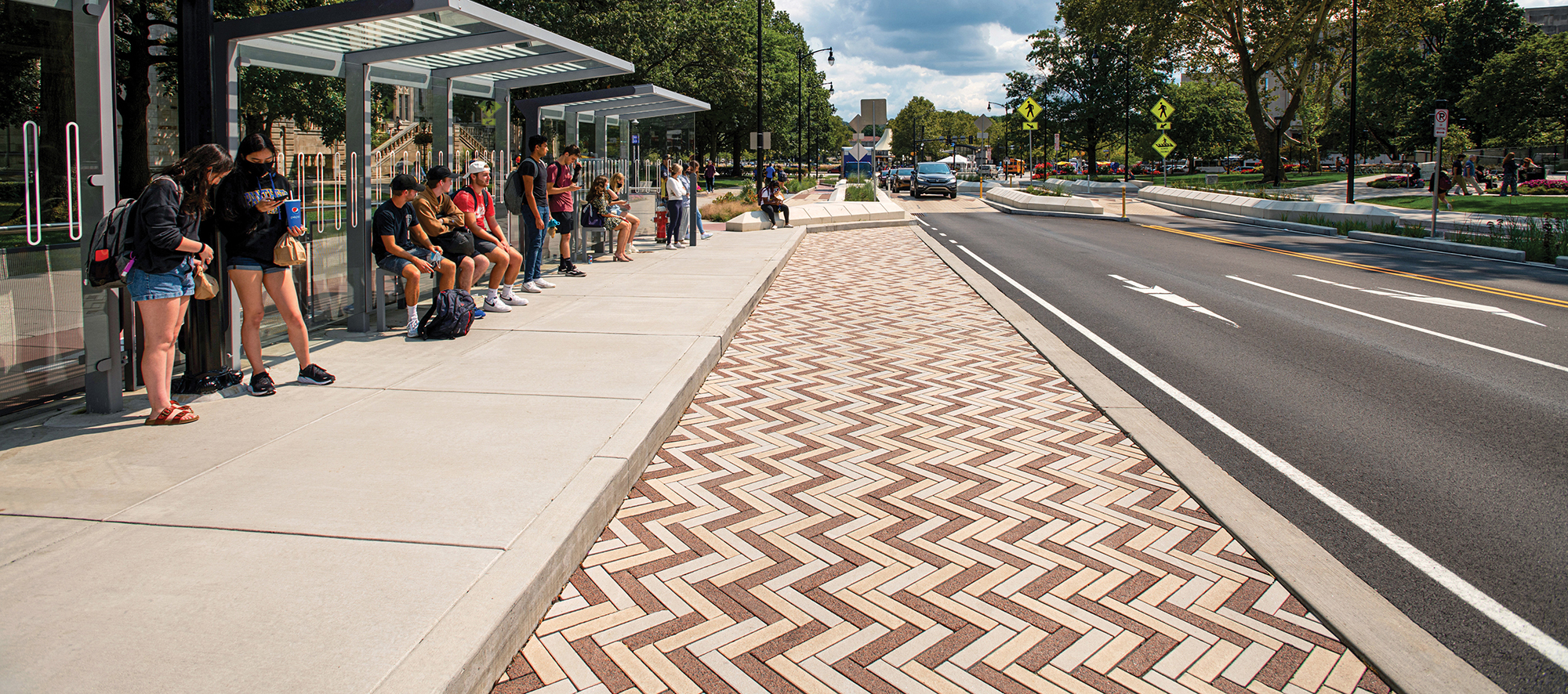 Students in summer clothing wait under bus shelters on a tree-lined road partially paved in Eco-Promenade in a colorful herringbone pattern.