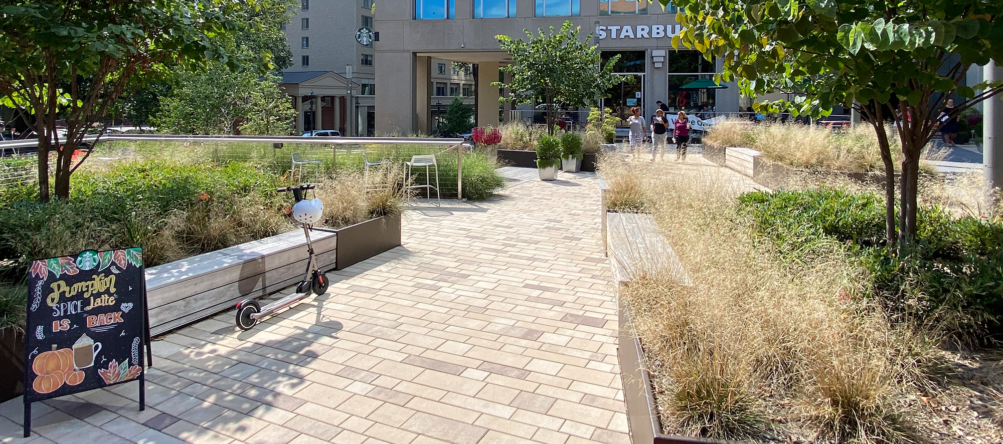 Three women walk out of a Starbucks onto a garden-lined pedestrian path made with Artline rectangular pavers in a mix of warm colors.