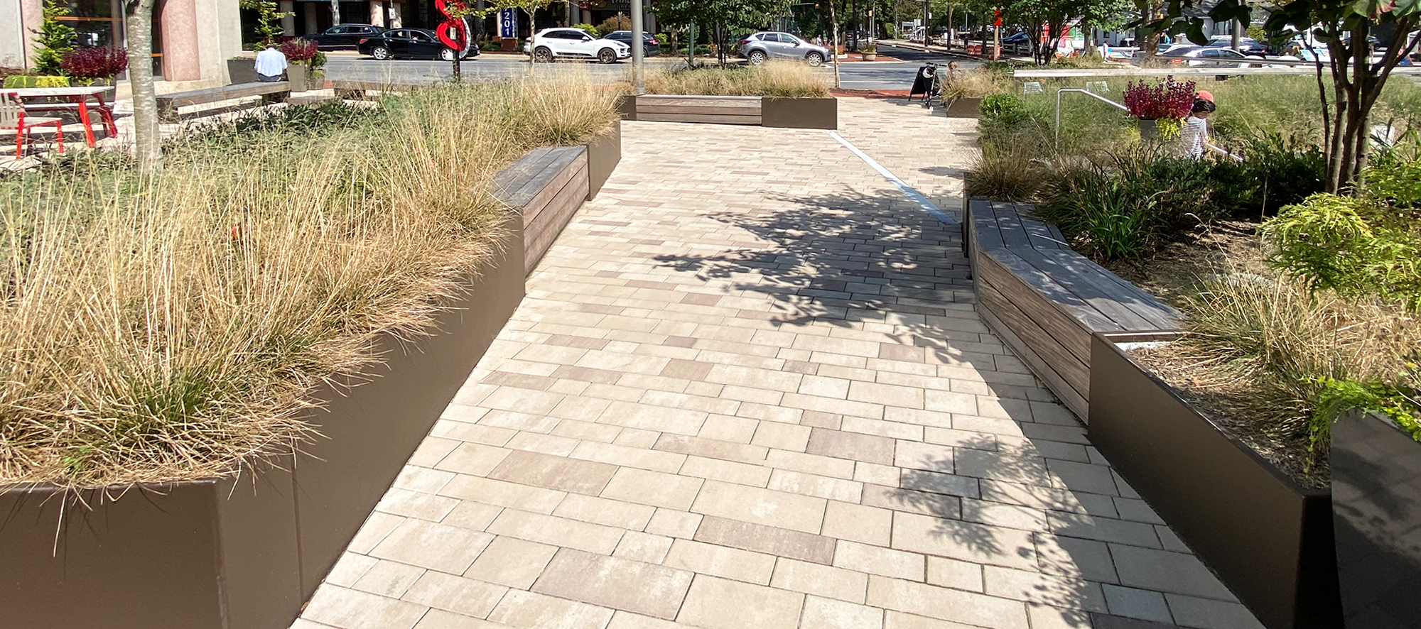A pedestrian plaza made with Unilock Artline rectangular pavers in a mix of warm colors, and raised gardens lead to the street.