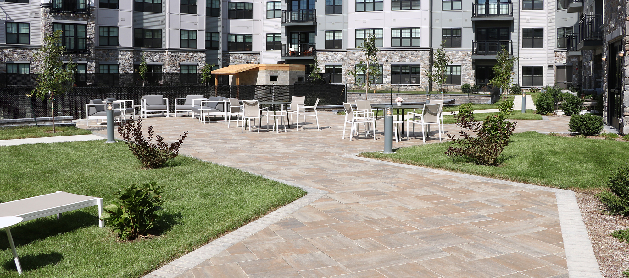 Outdoor gathering space in front of a grey building with ample seating, over brown Bristol Valley pavers lined with shrubs and grass.
