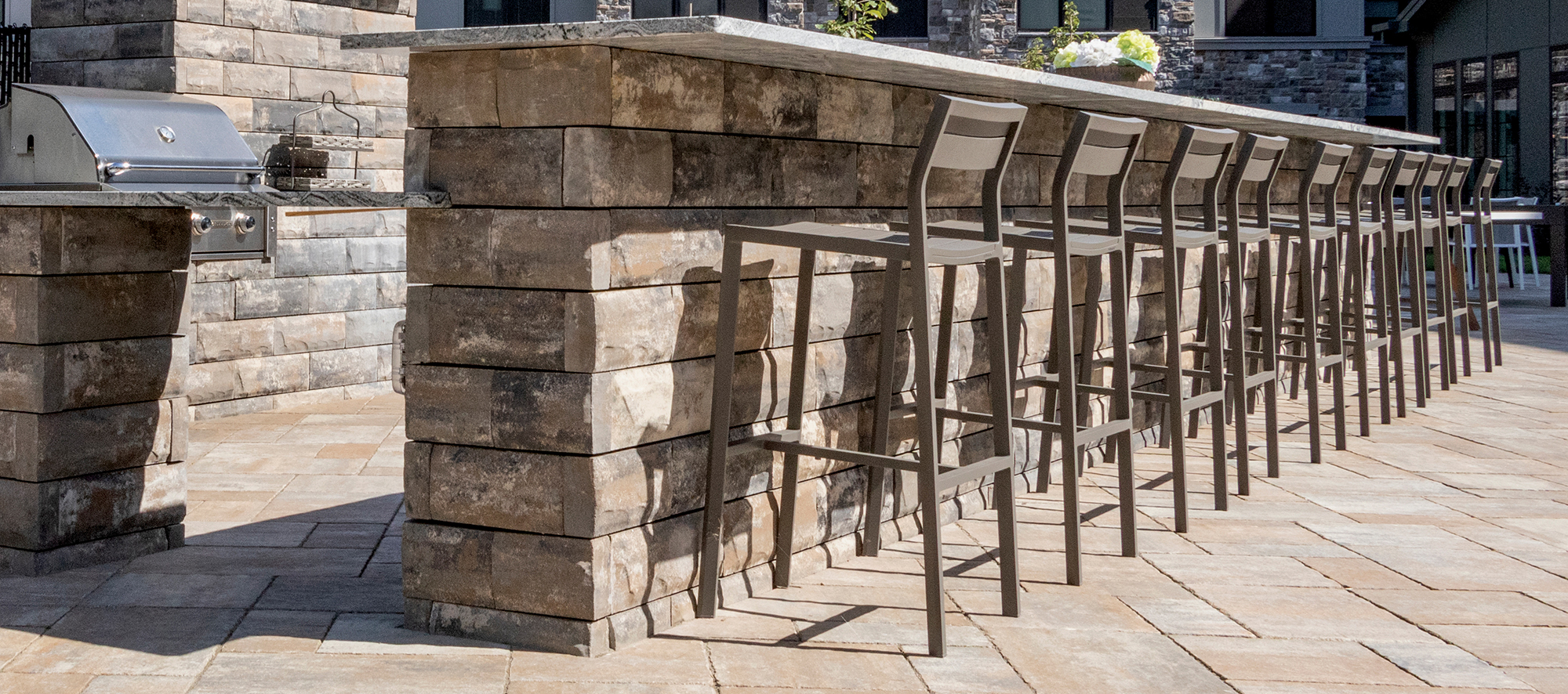 Bar stools neatly line a separate U-Cara bar island, with an adjacent outdoor barbecue on a paver bed of Bristol Valley brown pavers.