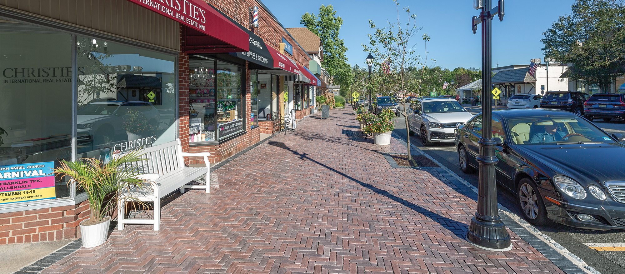 A raised sidewalk with warm tones of Copthorne pavers in a herringbone pattern has contrasting lines of black pavers on the sides.