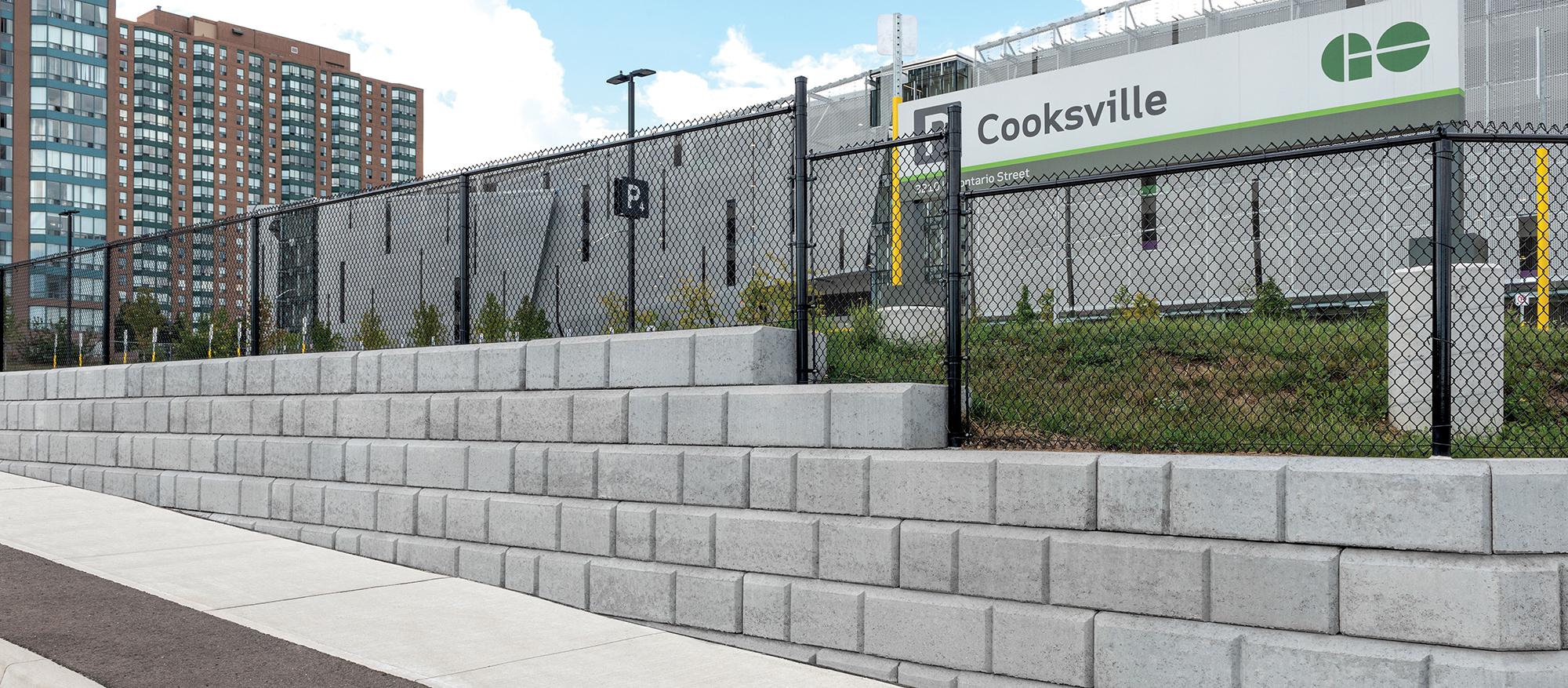 A retaining wall built with grey Dura-Hold blocks borders the perimeter of Cooksville Go station, complete with a black chain-link fence.