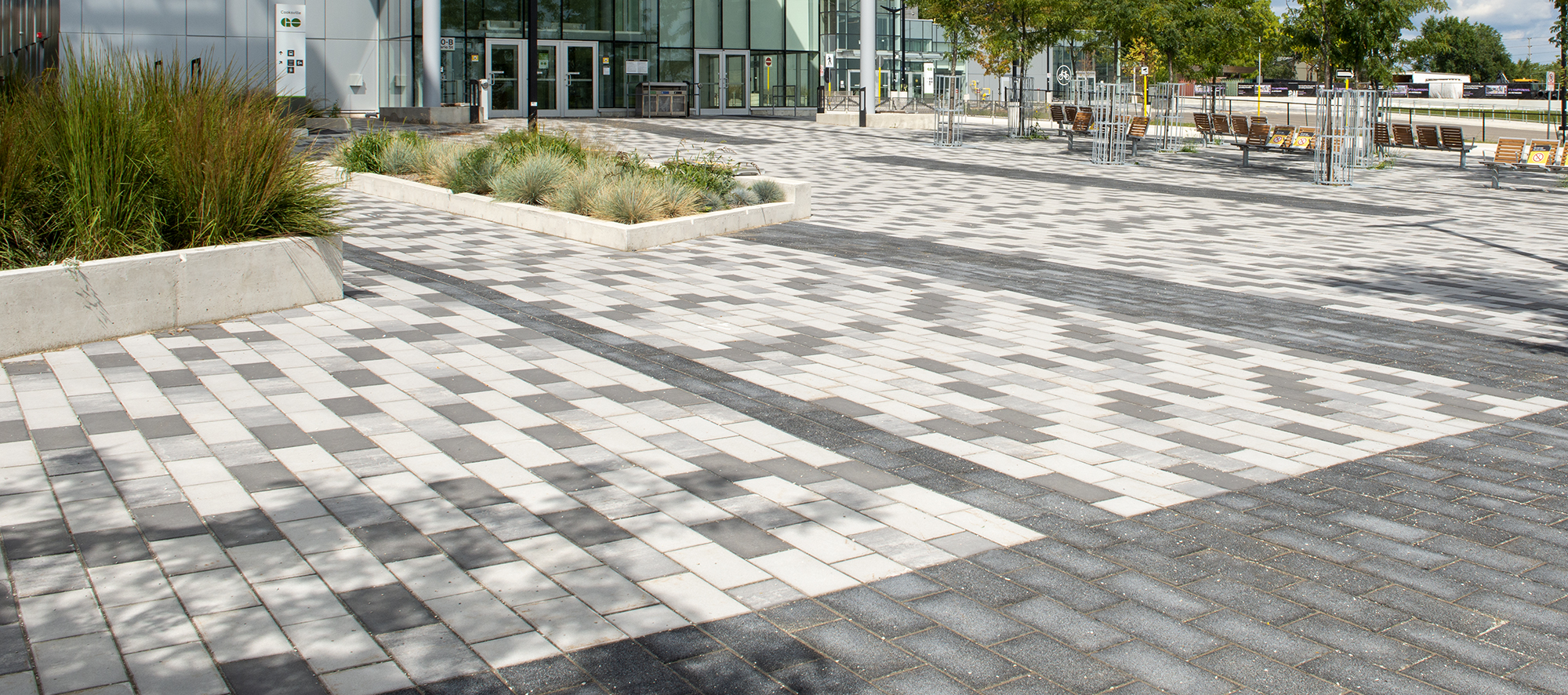 This expansive plaza features multiple park benches and natural greenery, combined with a paver bed of tri-colored Promenade Plank pavers.