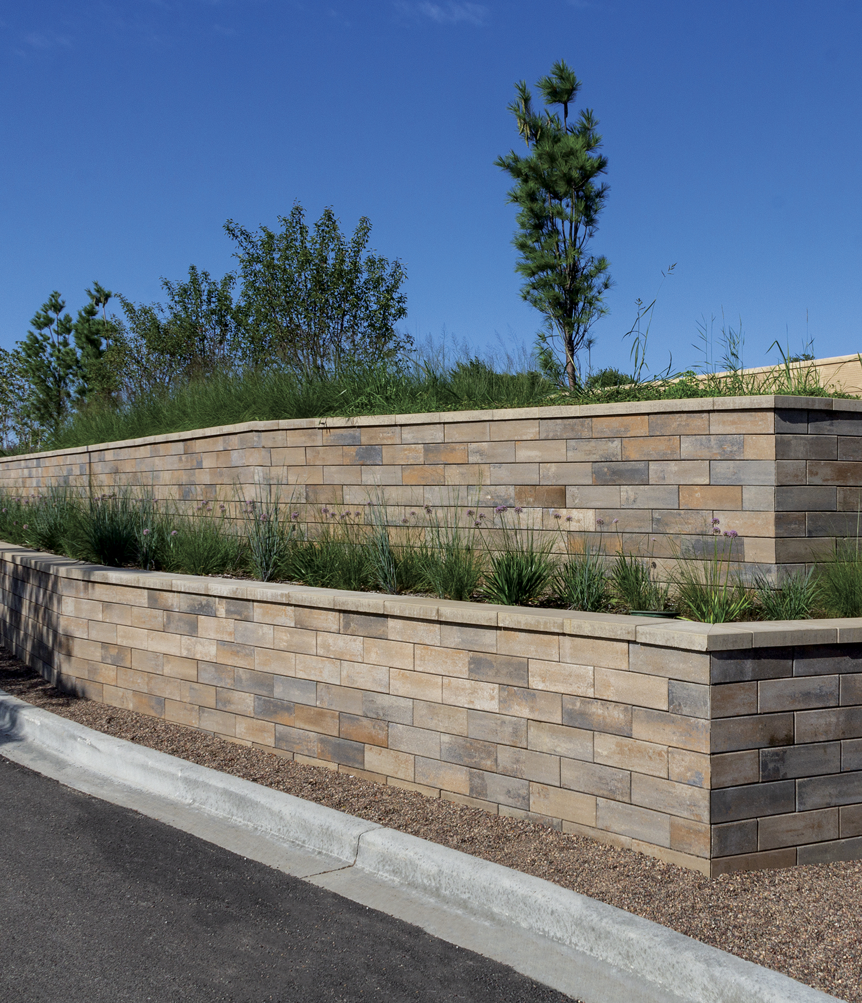 Beside a road is the side and front of a U-Cara wall with two levels and landscaping and trees in it.